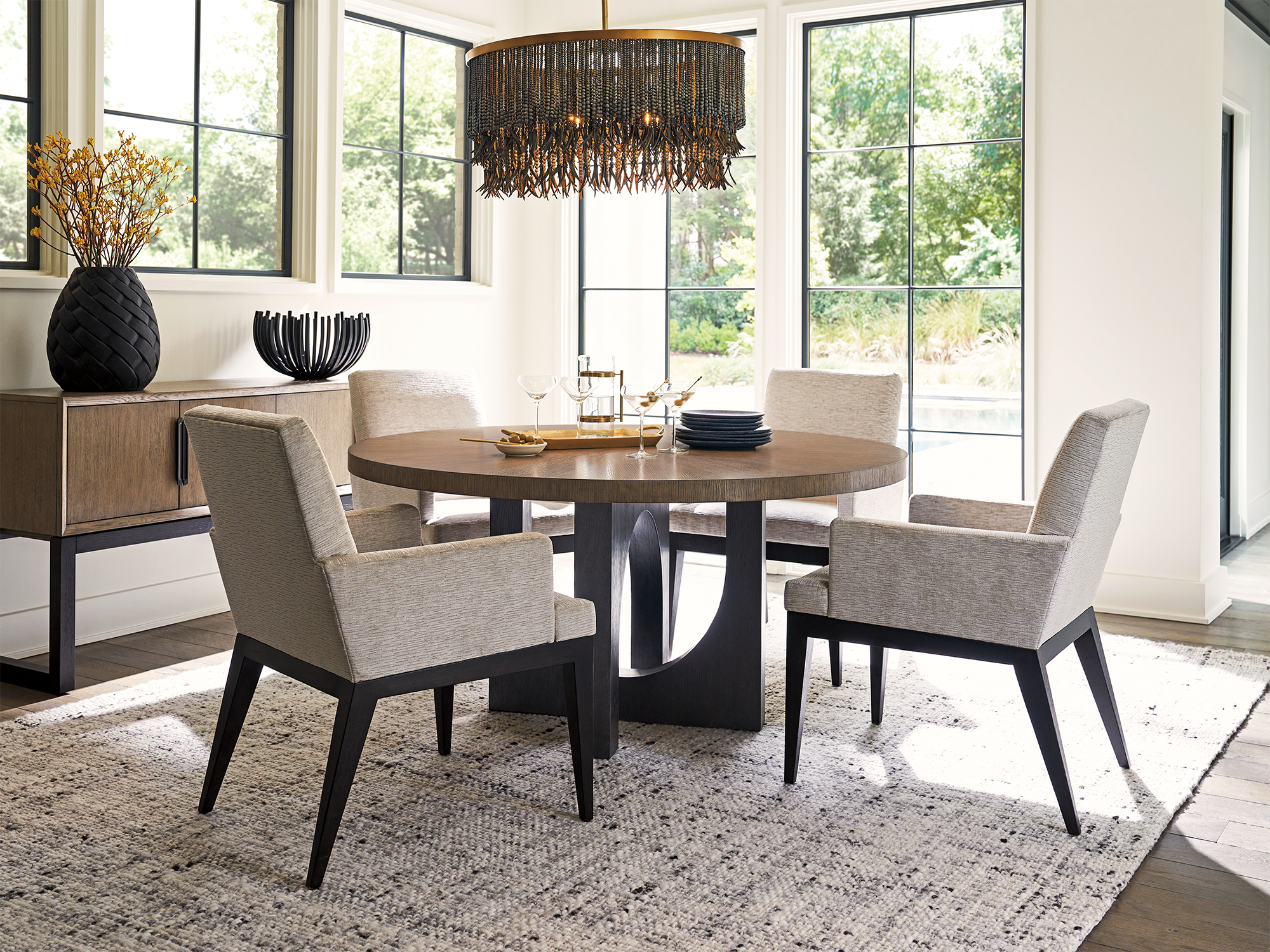 Lexington Zanzibar — global modern dining room featuring a round oak table with a sculptural black pedestal base, paired with gray upholstered armchairs and a tiered black bead chandelier.