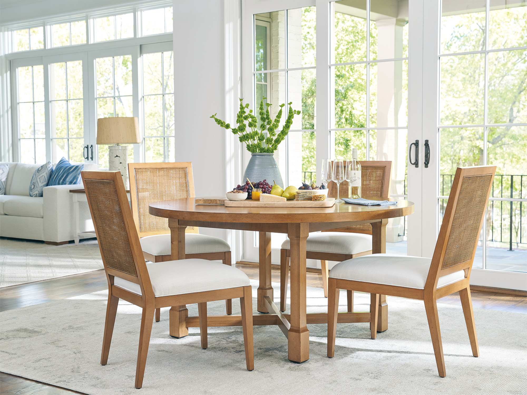 Barclay Butera for Lexington — Laguna collection coastal dining room featuring a round honey-toned oak table, cane-back dining chairs with white upholstered seats, and a bright, window-filled backdrop.