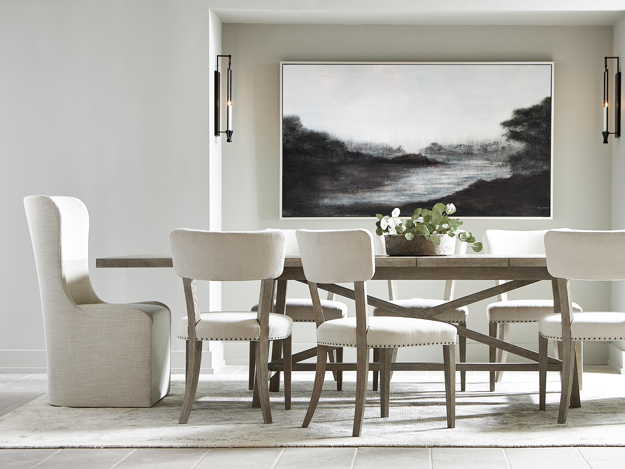 Bernhardt Albion — modern dining room featuring a long oak trestle table with an X-base design, paired with cream upholstered host and side chairs with nailhead trim on a area rug.