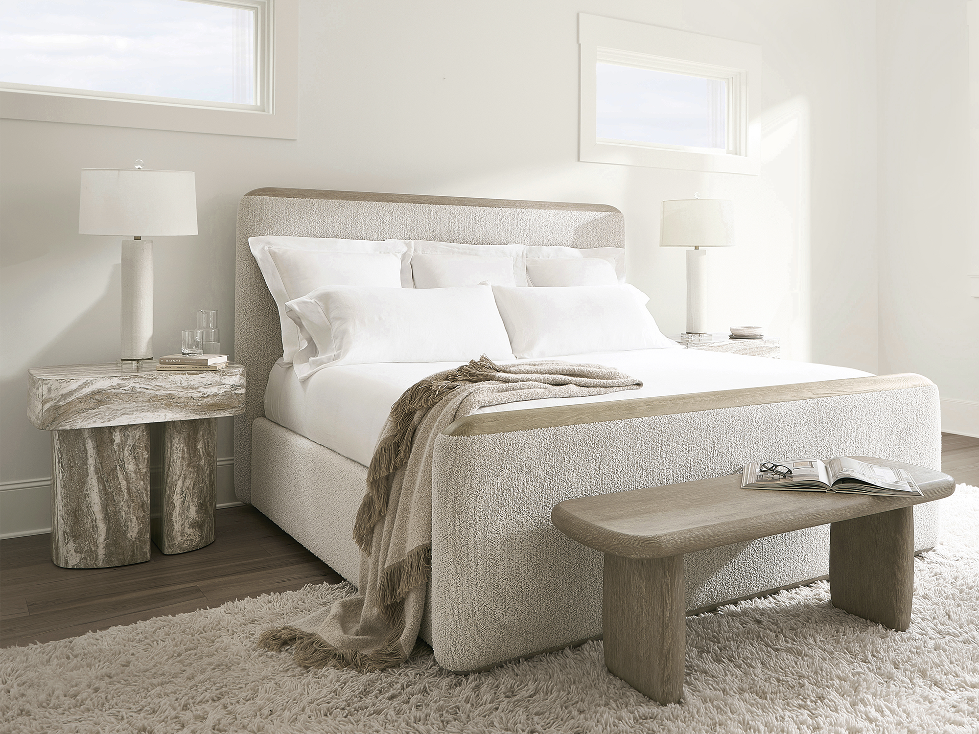 Bernhardt Arcadia — serene modern bedroom featuring a cream upholstered panel bed with woodgrain-textured cap rails in a Clay finish, a matching oak bench, and a totemic faux-stone nightstand in Kalta.