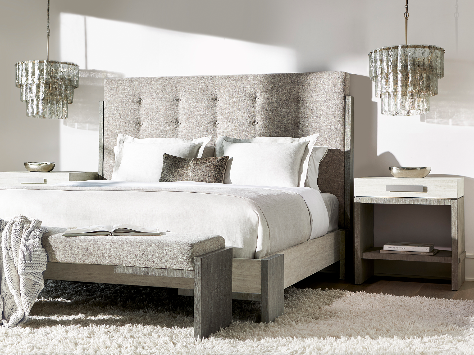 Bernhardt Foundations — modern luxury bedroom featuring a light gray tufted upholstered bed with a tall headboard, paired with matching two-tone nightstands, a cushioned bench, and tiered glass chandeliers.
