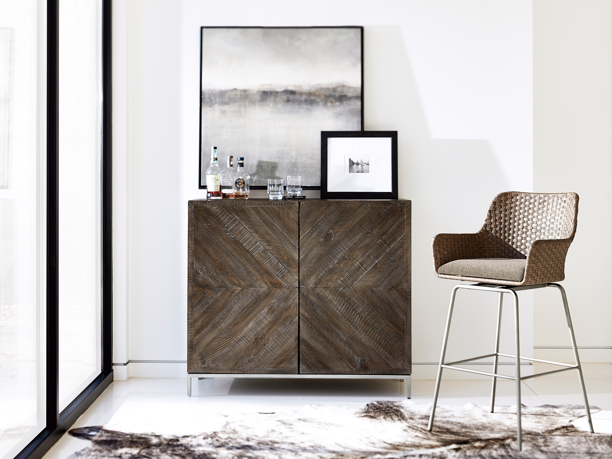 Bernhardt Loft Logan Square — contemporary bar or dining area featuring a dark wood cabinet with a distinctive chevron-patterned facade, paired with a woven high-back swivel barstool and a landscape painting, set on a natural hide rug.