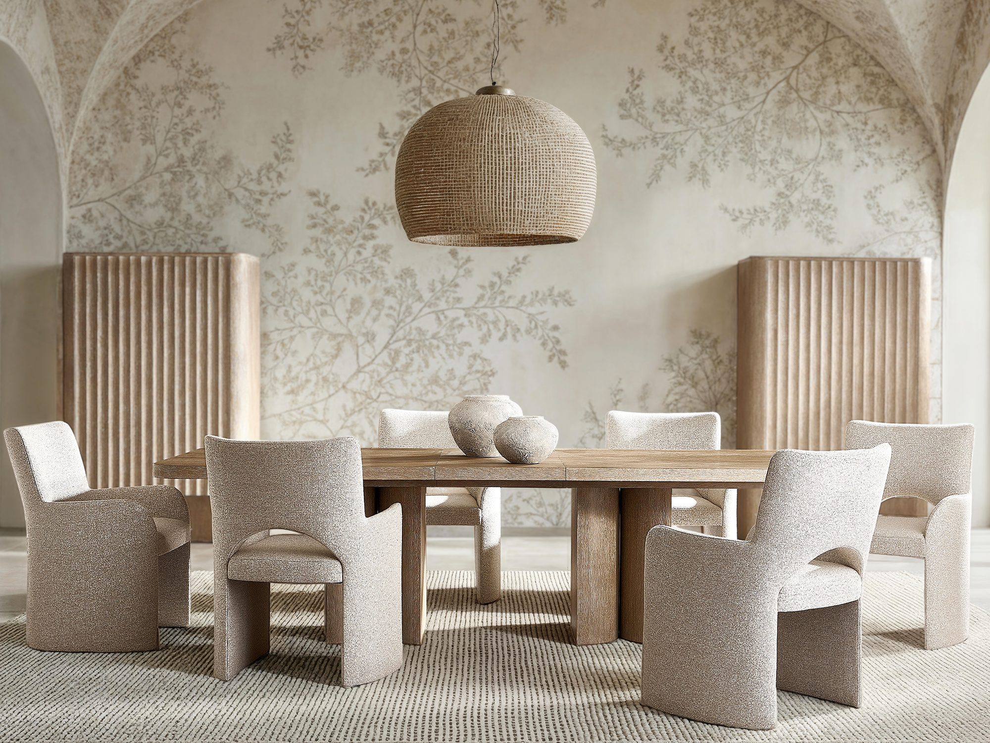 Bernhardt Loggia — Mediterranean-inspired dining room featuring an oak trestle table with half-cylinder pedestal legs, sculptural bouclé armchairs, and fluted arched cabinets against a botanical fresco wall.