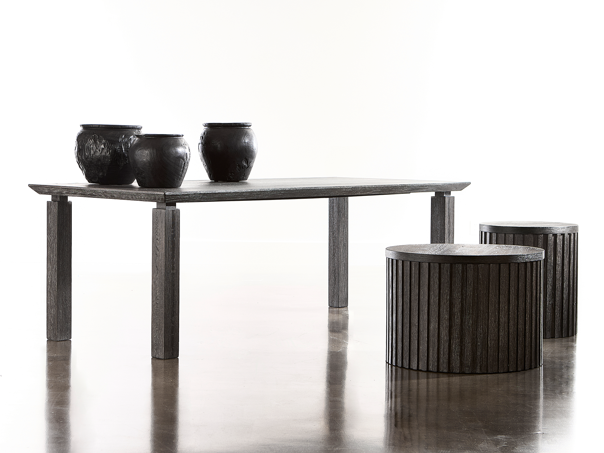Bernhardt Menton — minimalist dining or workspace featuring a dark charcoal-finished rectangular table with block legs, paired with two matching cylindrical fluted stools and large textured black ceramic vessels on a high-sheen concrete floor.