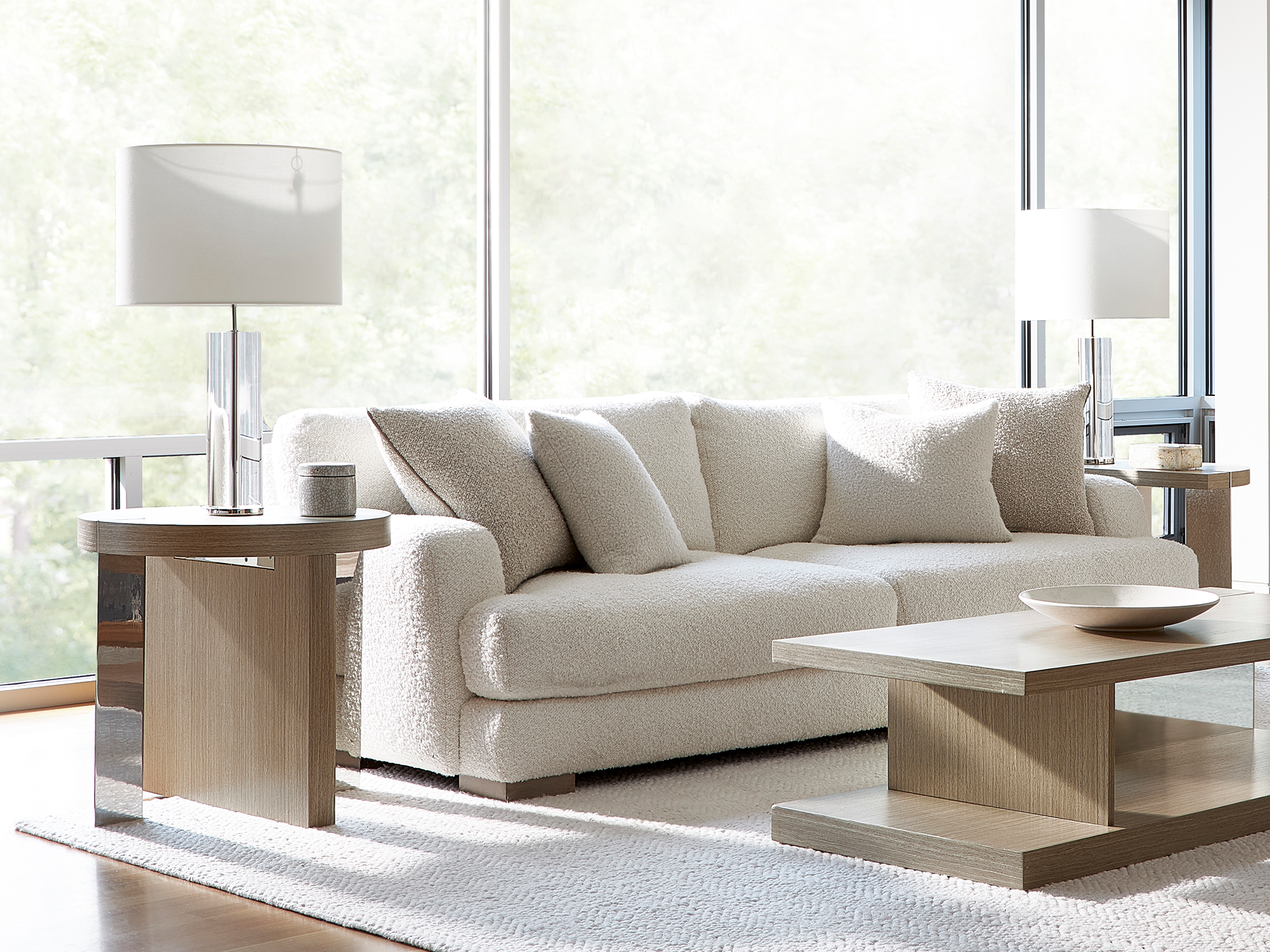 Bernhardt Modulum — ultra-modern living room featuring a plush cream bouclé sofa with oversized pillows, paired with light oak-finished tiered coffee and end tables, accented by sleek glass lamps in a minimalist, sun-lit space.