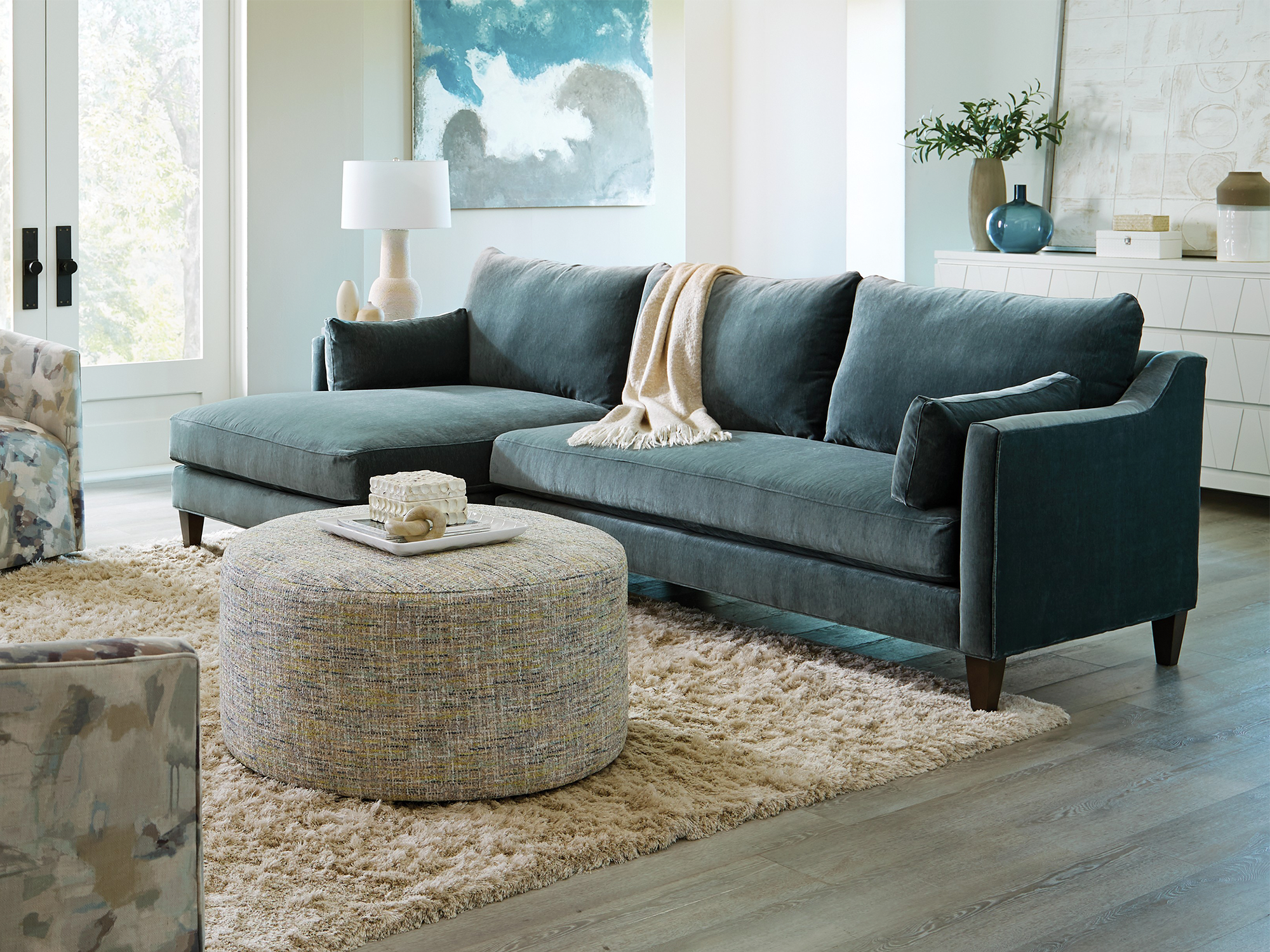 Craftmaster CM Modern — contemporary living room featuring a sleek teal velvet sectional with a left-facing chaise, a large round textured fabric ottoman, and watercolor-patterned accent chairs on a plush shag rug.
