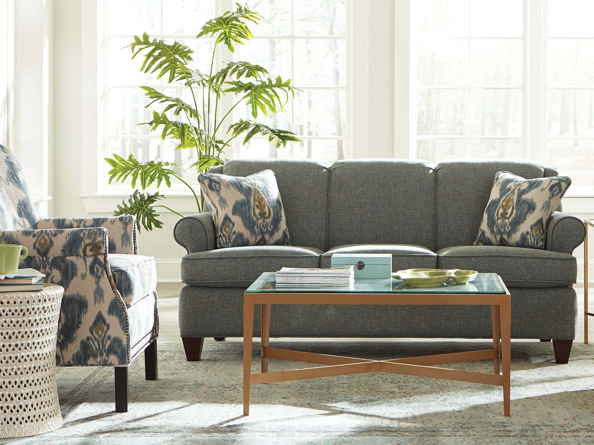 Craftmaster Essentials — traditional living room featuring a slate blue textured sofa with rolled arms, an ikat-patterned accent chair with nailhead trim, and a glass-top coffee table with a gold X-base frame in a sun-drenched space.