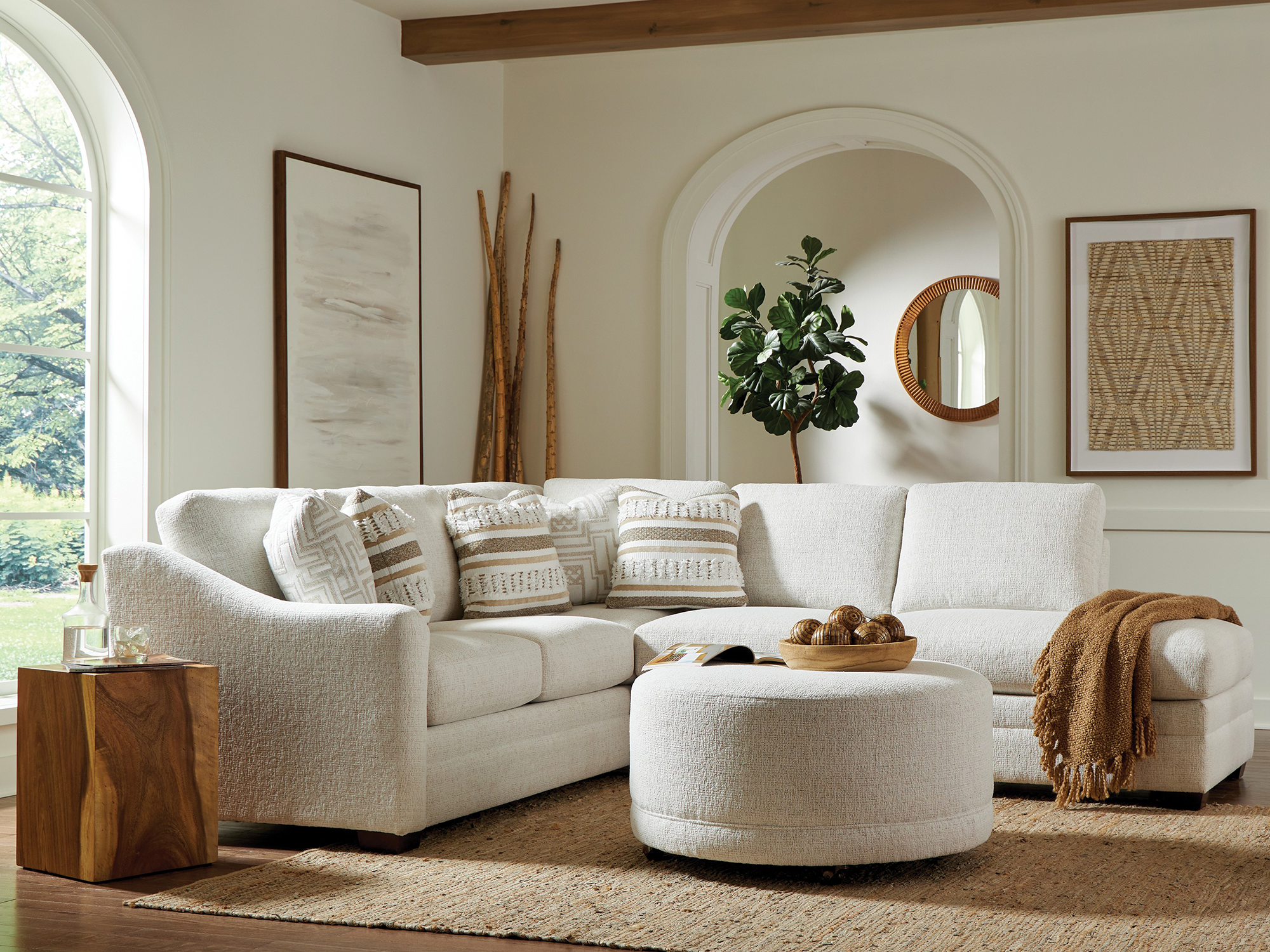 Craftmaster Design Options — modern transitional sectional featuring a cream textured fabric, a matching round cocktail ottoman, and neutral-toned geometric accent pillows, paired with a solid wood cube end table in an organic, light-filled living space.