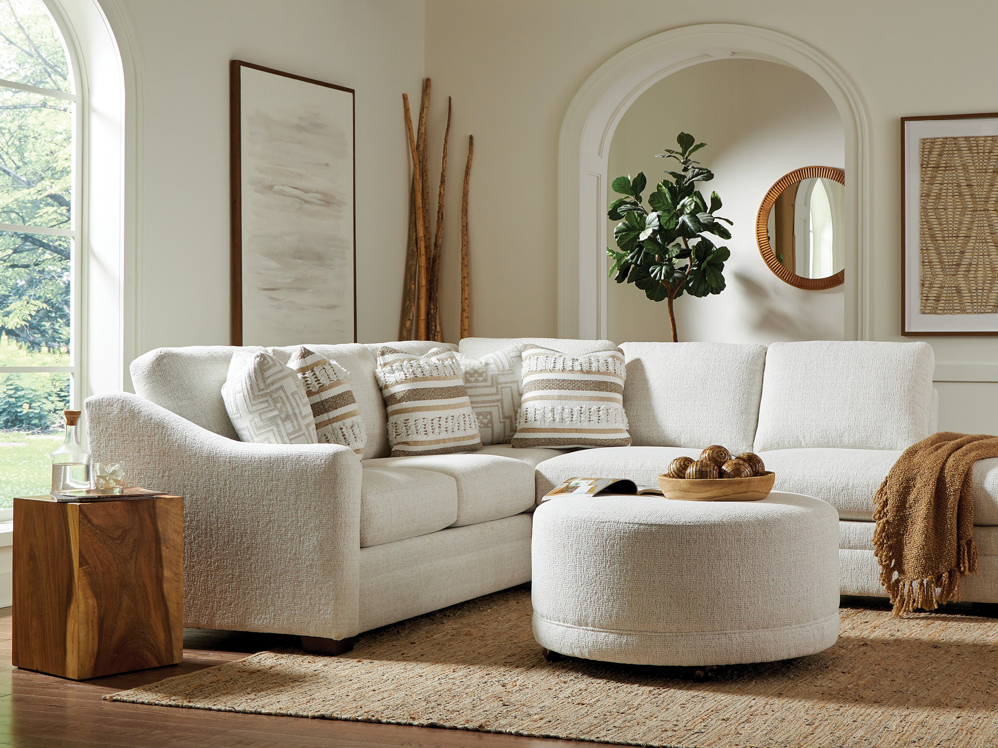 Craftmaster Furniture — transitional living room featuring a cream textured fabric sectional with track arms, a matching oversized round cocktail ottoman, and a solid wood block end table with visible natural grain.