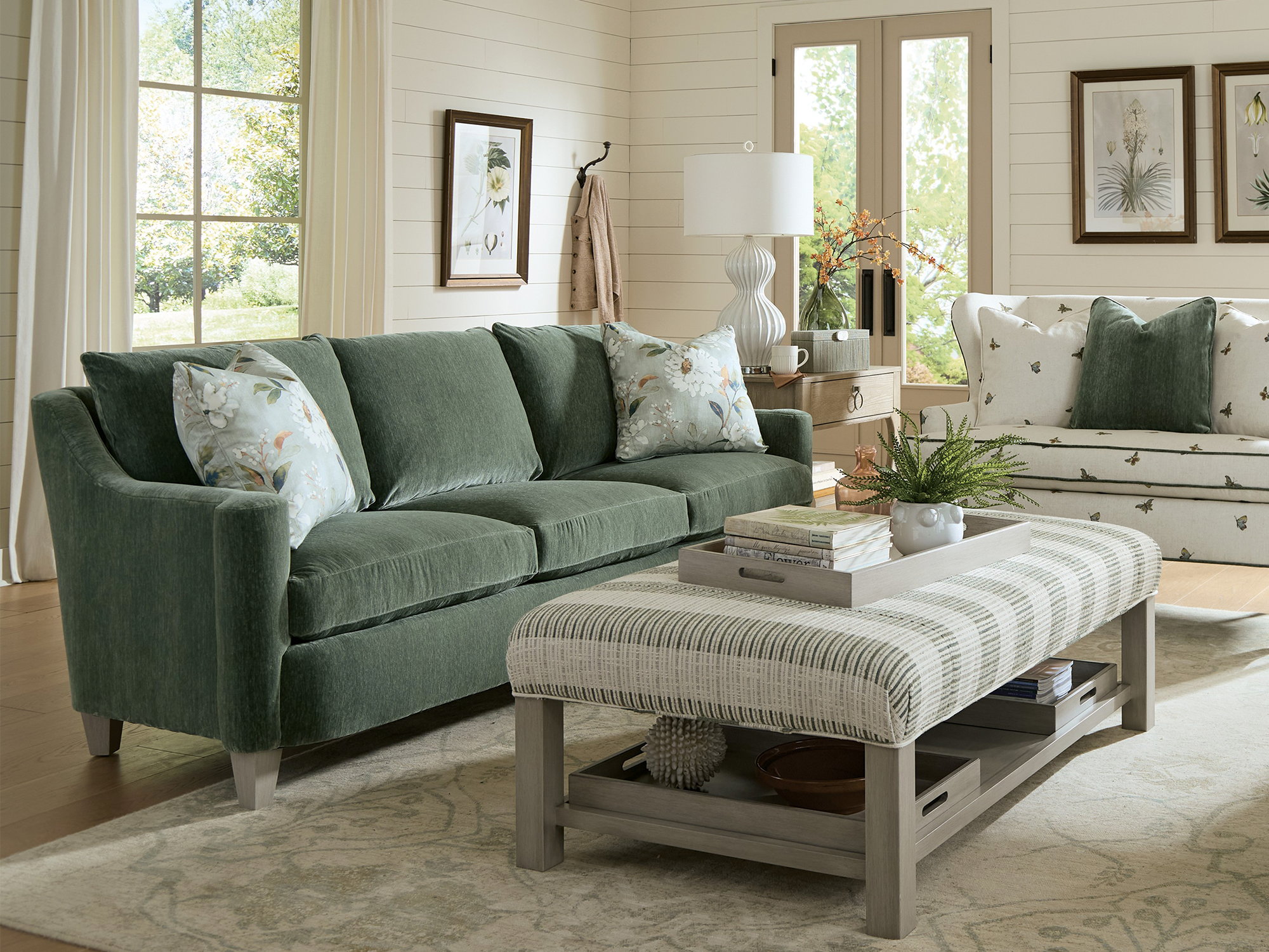 Craftmaster Paula Deen Home Collection — cottage-style living room featuring a deep green velvet sofa with floral accent pillows, a striped upholstered cocktail ottoman with a wood base, and a patterned wingback loveseat in a bright, airy sunroom.