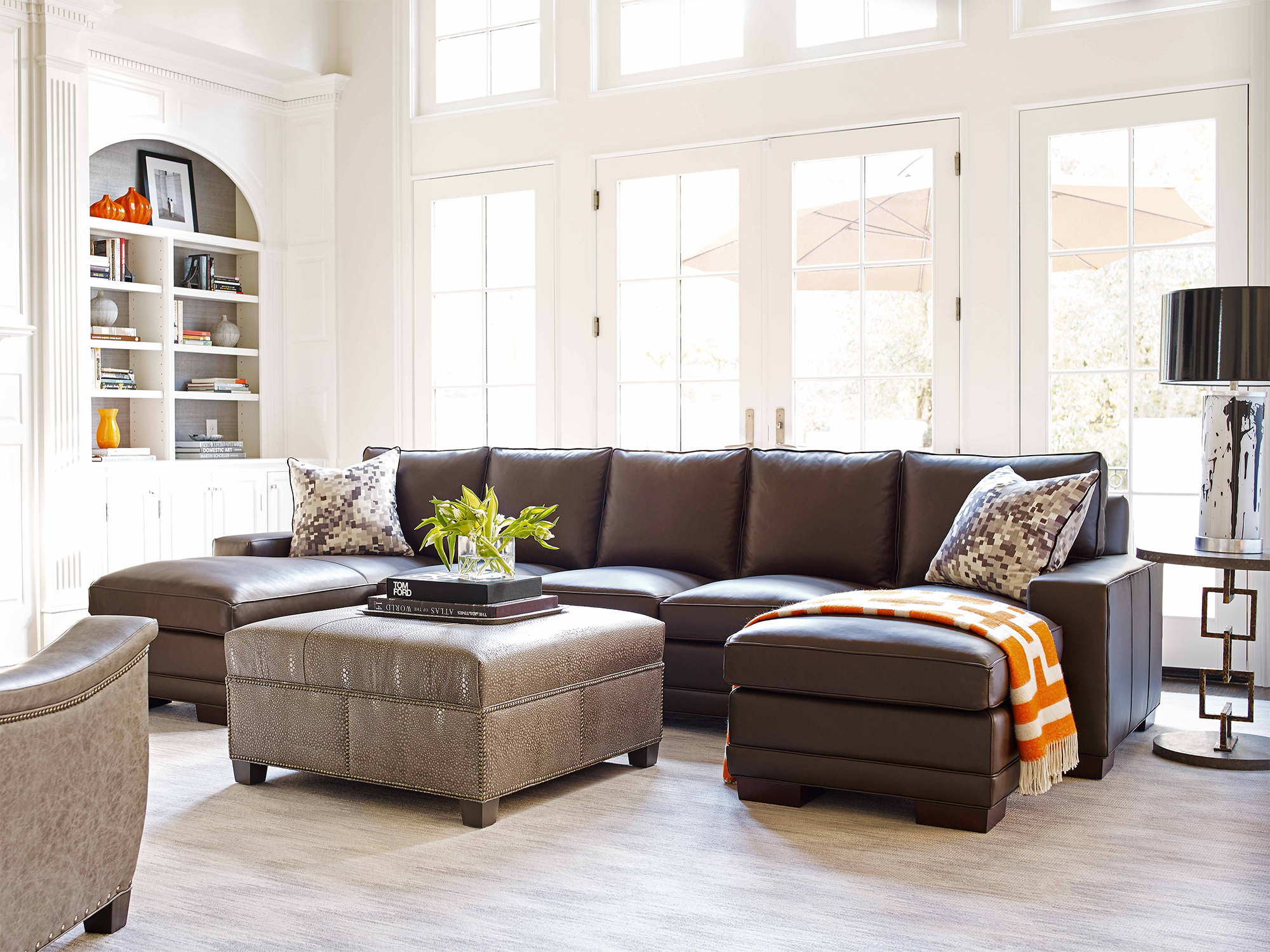 Hancock & Moore — modern living room featuring a dark chocolate leather sectional with a double-chaise design, paired with a large square embossed-leather cocktail ottoman with nailhead trim and an orange geometric accent throw.