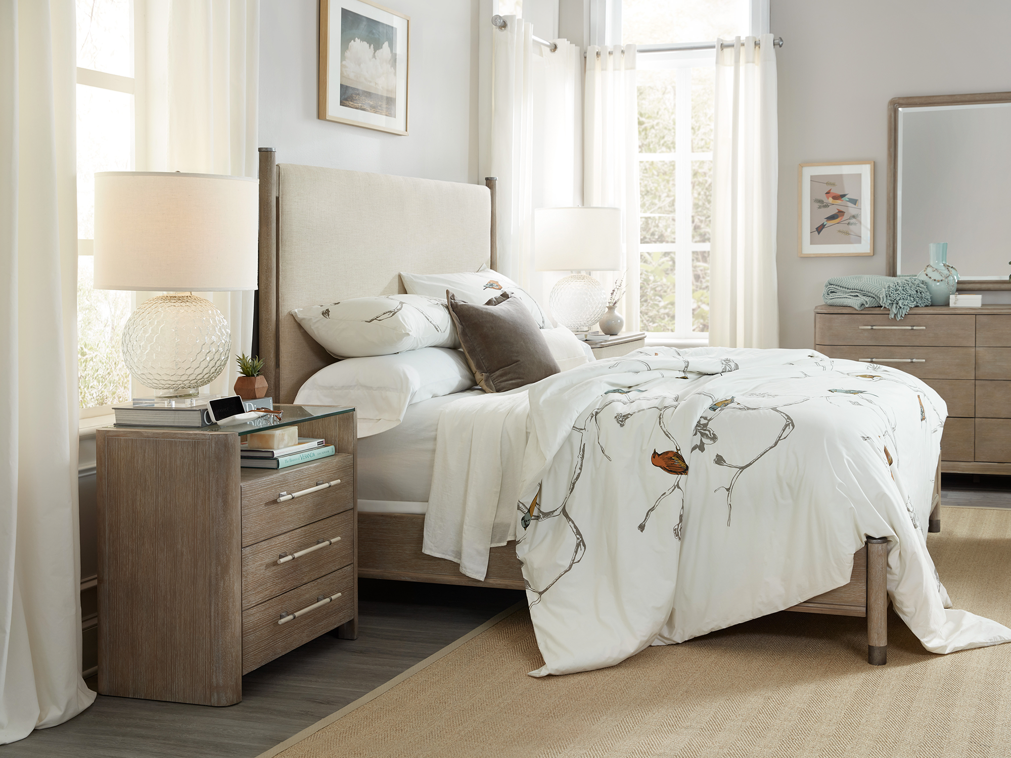 Hooker Furniture Affinity — serene transitional bedroom featuring a greige-washed panel bed with a cream upholstered headboard, a glass-top three-drawer nightstand, and bird-print botanical bedding.