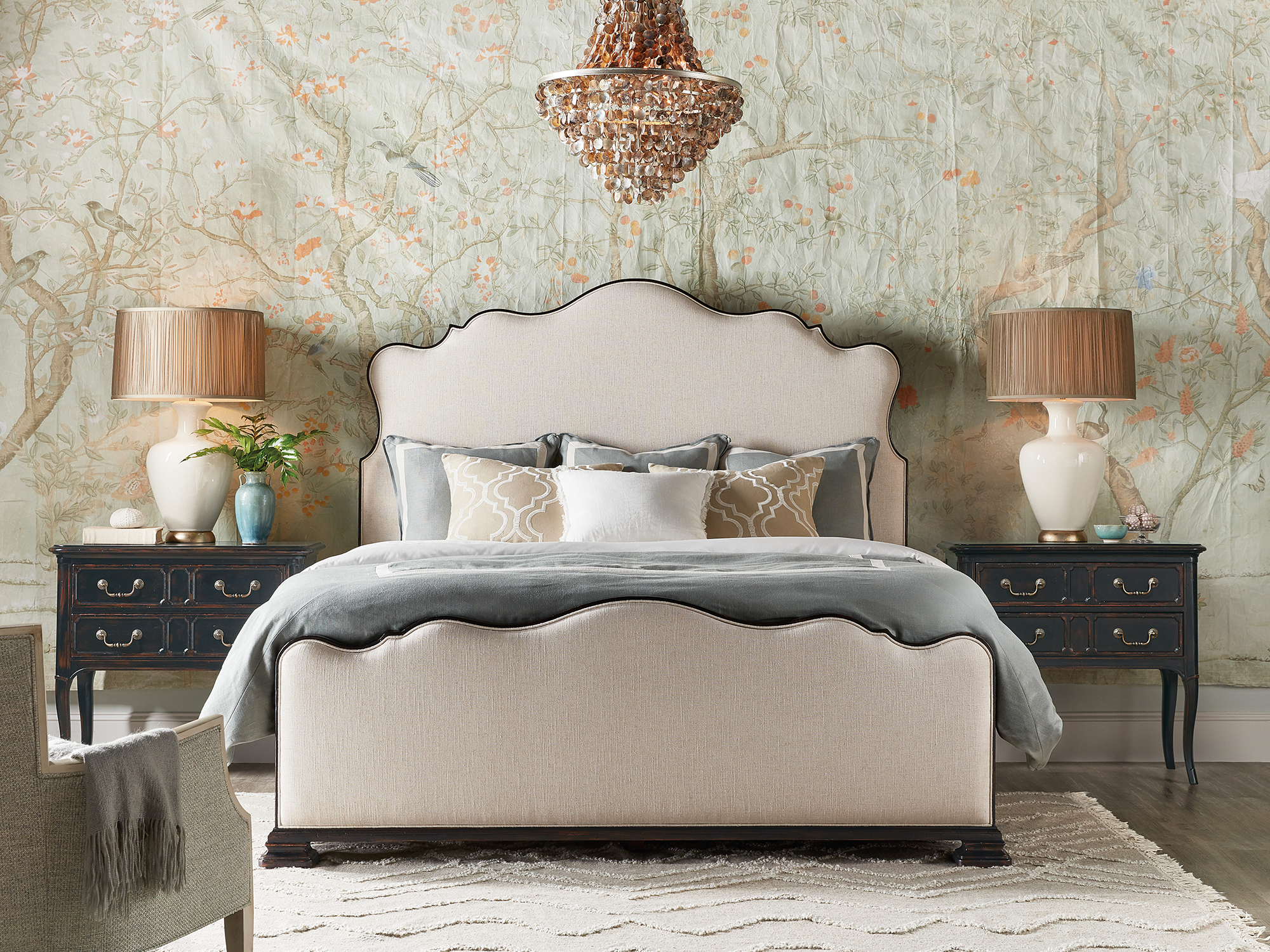 Hooker Furniture — Charleston collection traditional bedroom featuring an upholstered scalloped bed with dark wood trim, paired with black three-drawer nightstands and a beaded crystal chandelier against a floral chinoiserie wallpaper.
