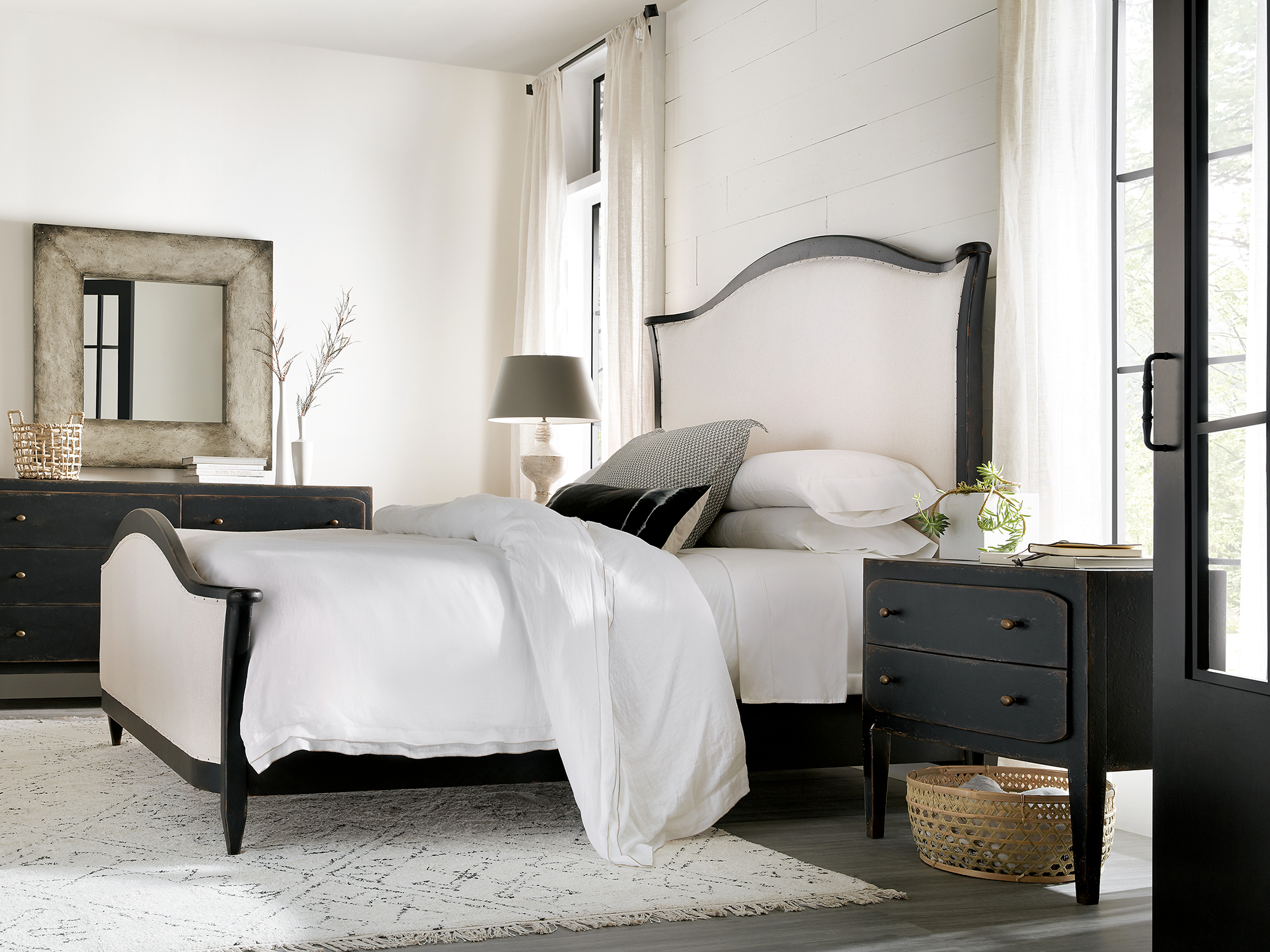 Hooker Furniture Ciao Bella — rustic chic bedroom featuring a black-distressed farmhouse bed with a cream upholstered headboard, a matching two-drawer nightstand, and a weathered silver leaf mirror.