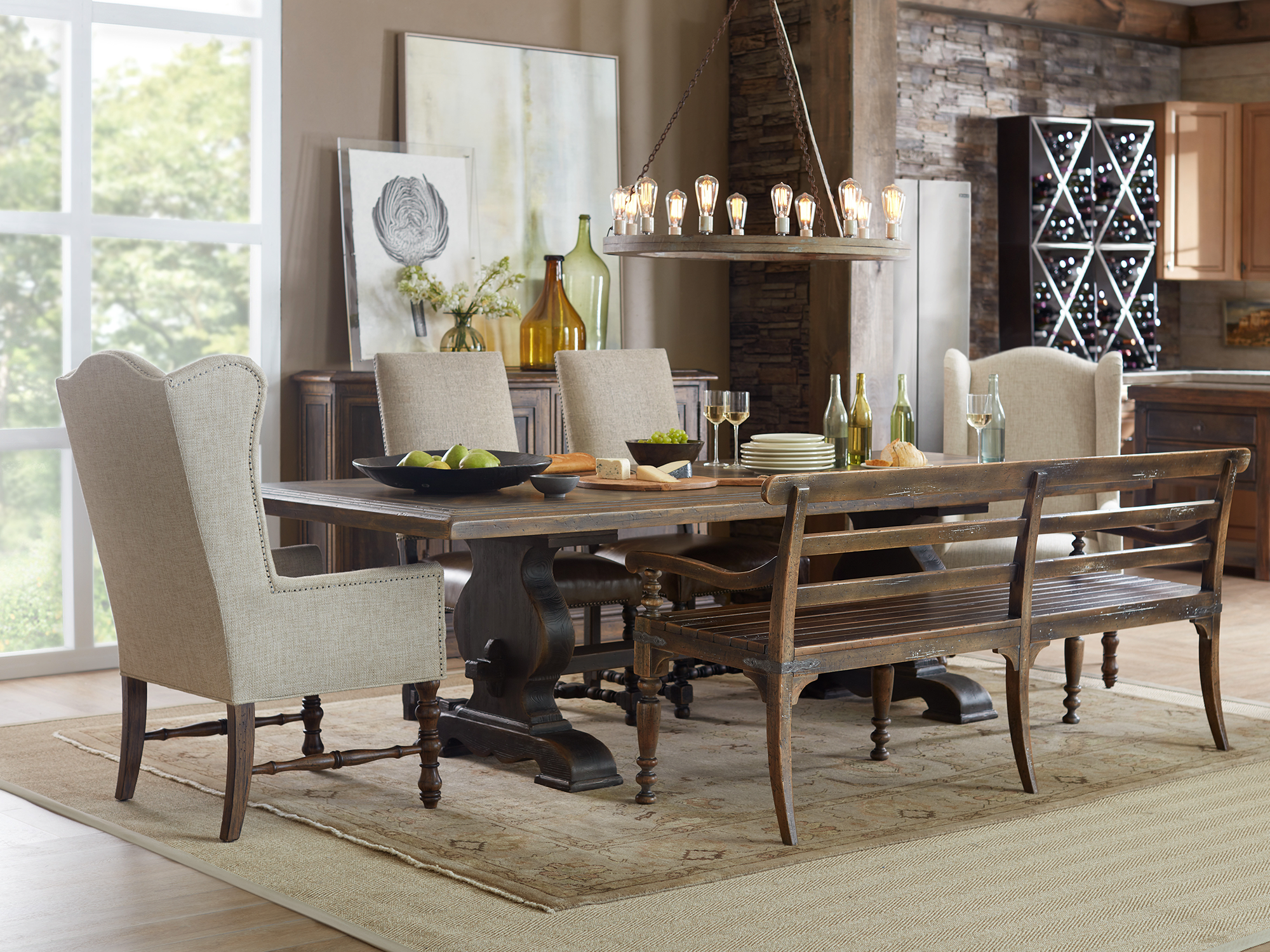 Hooker Furniture Hill Country — rustic refined dining room featuring a heavy distressed oak trestle table, a vintage-style wooden bench, and linen wingback host chairs under a wagon wheel chandelier.