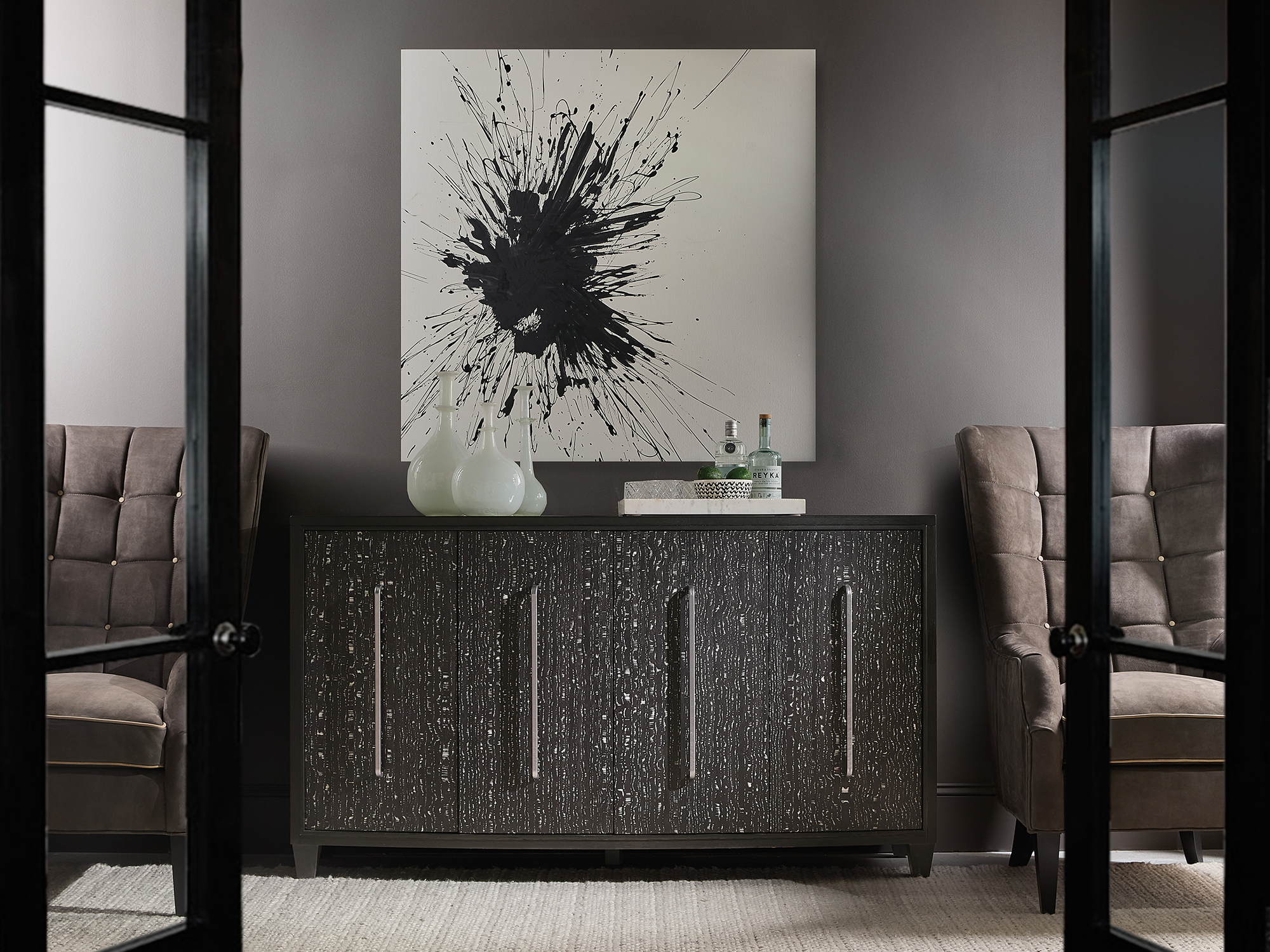 Hooker Furniture Mélange — sophisticated modern vignette featuring a charcoal buffet with silver textured door fronts and oversized bar pulls, flanked by tufted velvet wingback chairs.