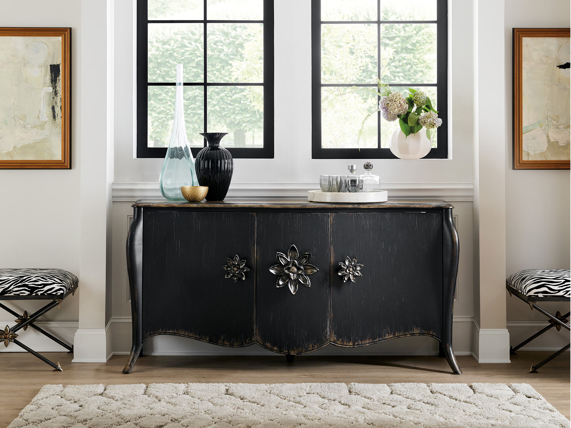 Hooker Furniture Sanctuary — dramatic glam vignette featuring a black-distressed bombe sideboard with oversized floral medallion hardware, flanked by zebra-print X-base stools.
