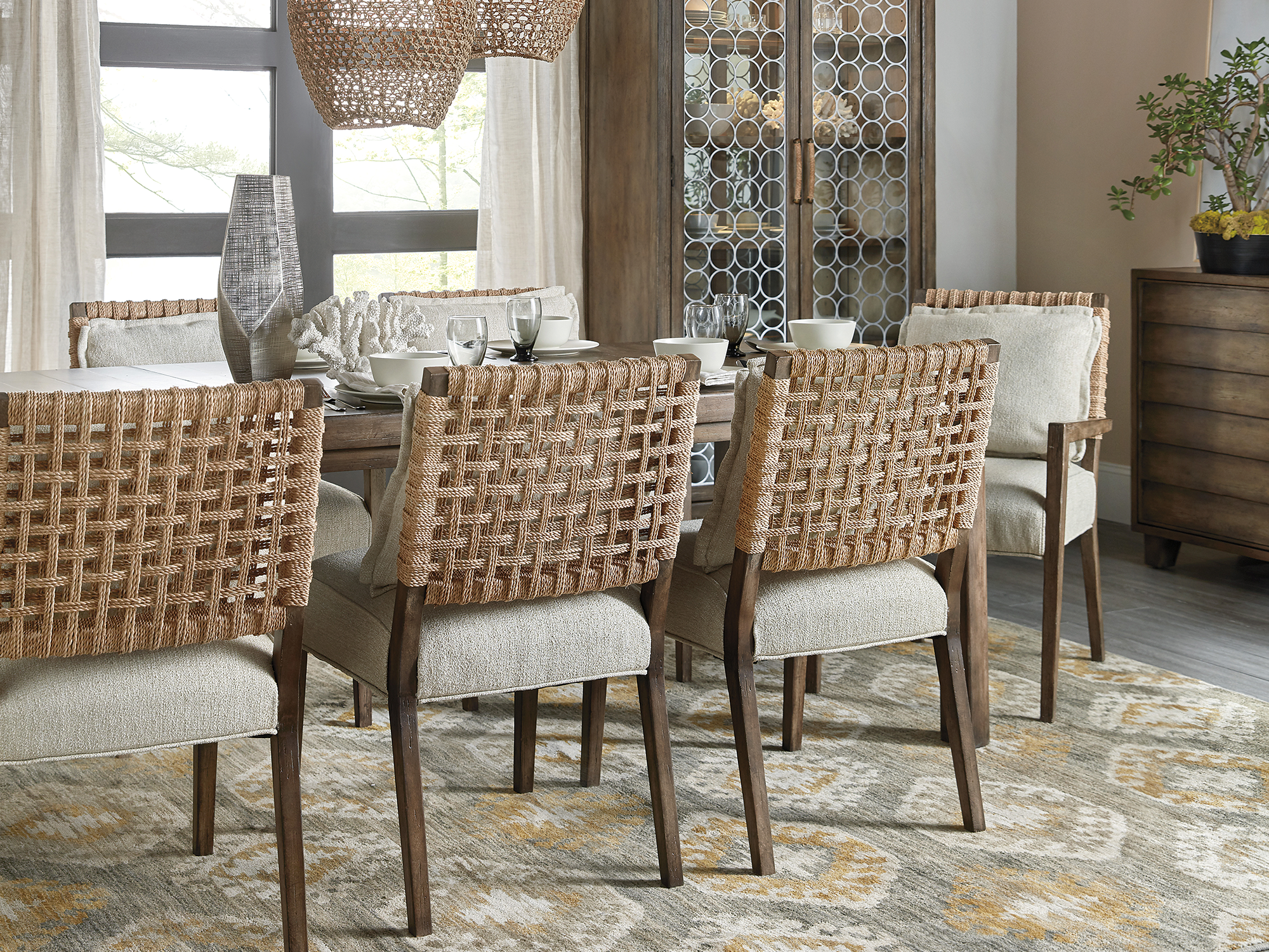 Hooker Furniture Sundance — organic coastal dining room featuring "woven-rope" back chairs, a distressed wood trestle table, and an architectural china cabinet with decorative circular fretwork.