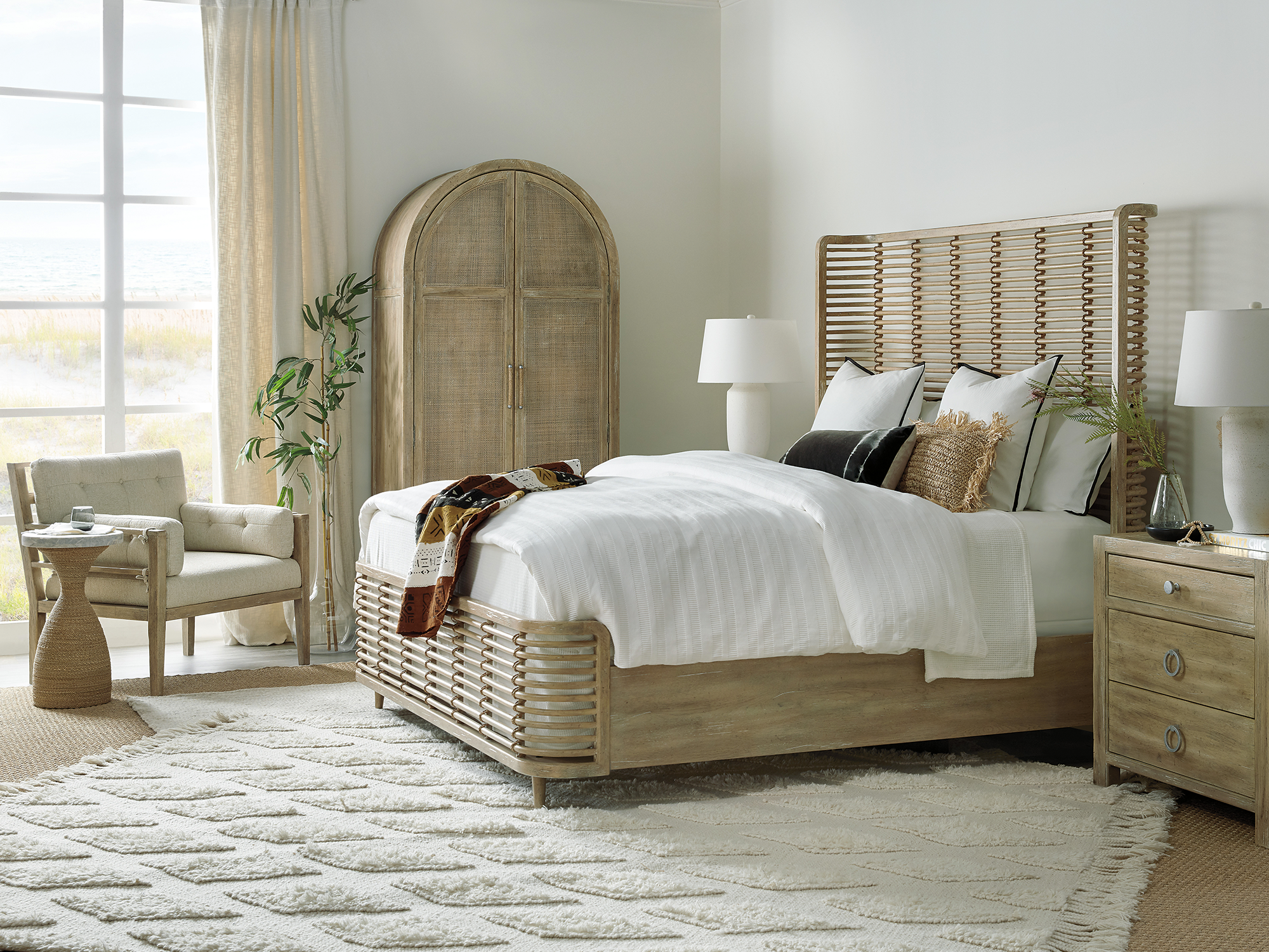 Hooker Furniture Surfrider — breezy coastal bedroom featuring a statement "split-rattan" bed, an arched cane-front armoire, and a weathered oak nightstand overlooking a beach dunes view.