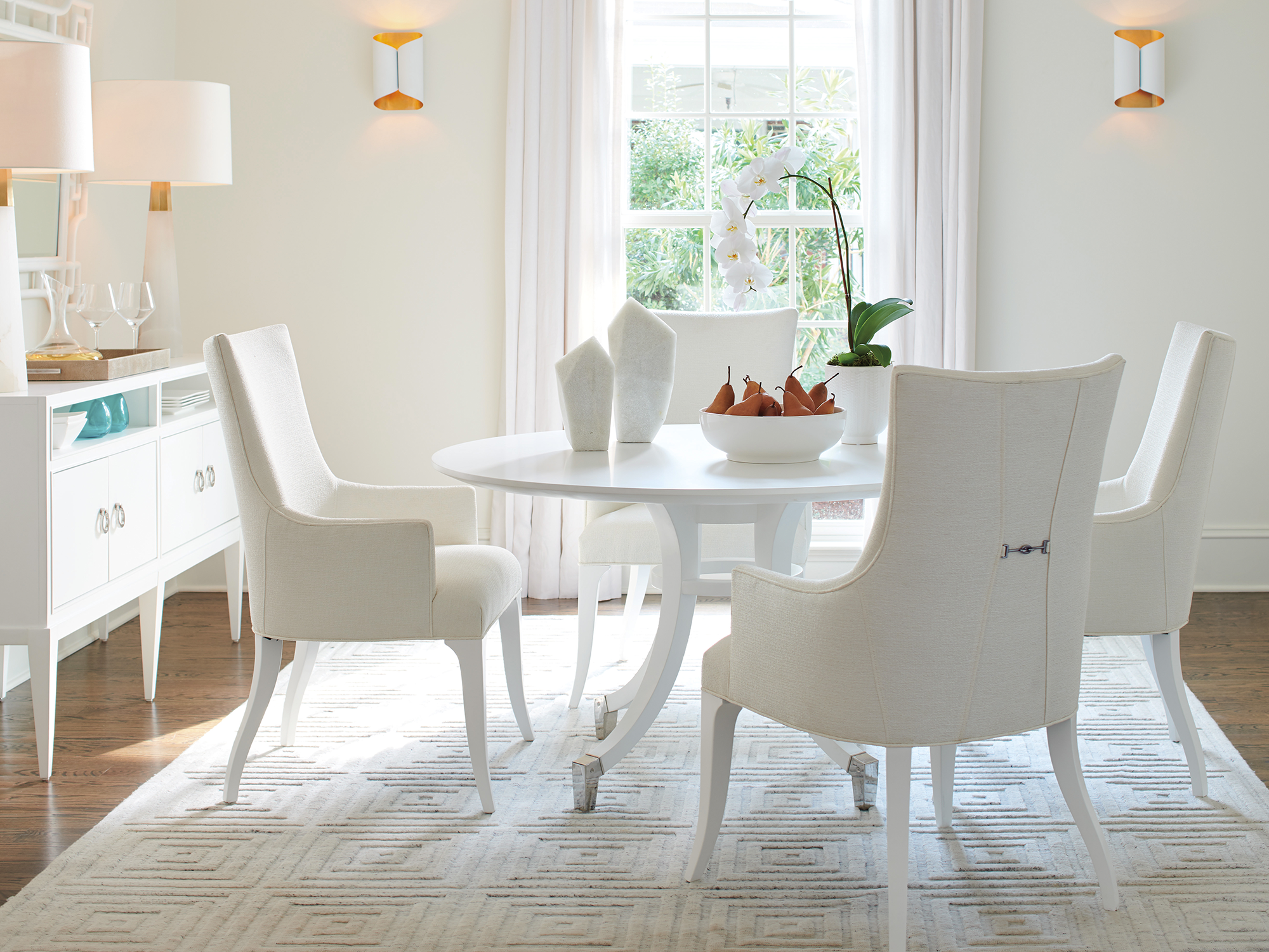 Lexington Avondale — Round white pedestal dining table and upholstered klismos-style armchairs with metal back details. This bright, monochromatic space features a matching white sideboard and geometric rug for a chic, contemporary aesthetic.