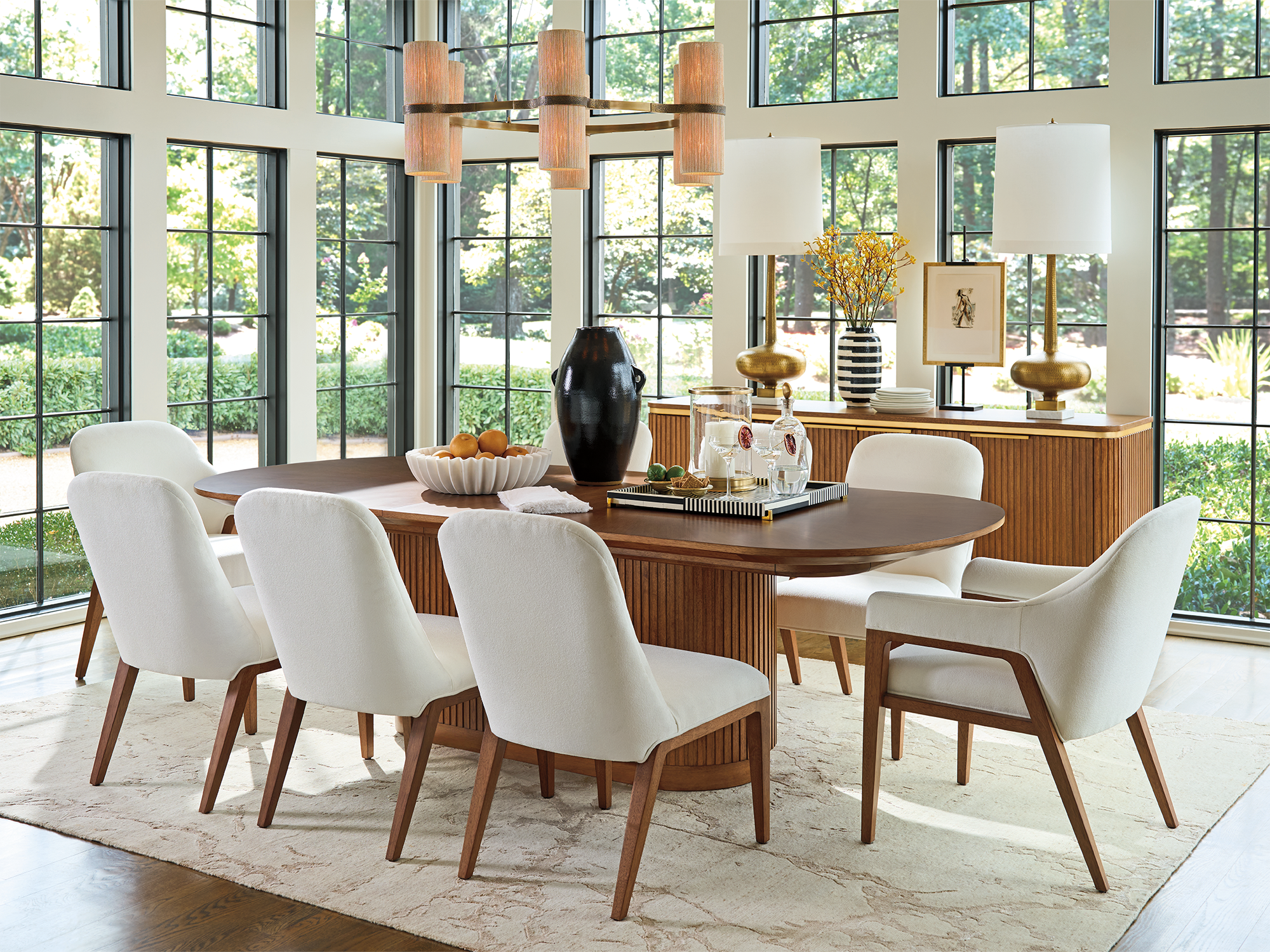 Lexington La Costa — modern organic dining room featuring an oval walnut table with a fluted pedestal base, cream sculptural dining chairs, and a matching fluted sideboard against a garden-view window wall.