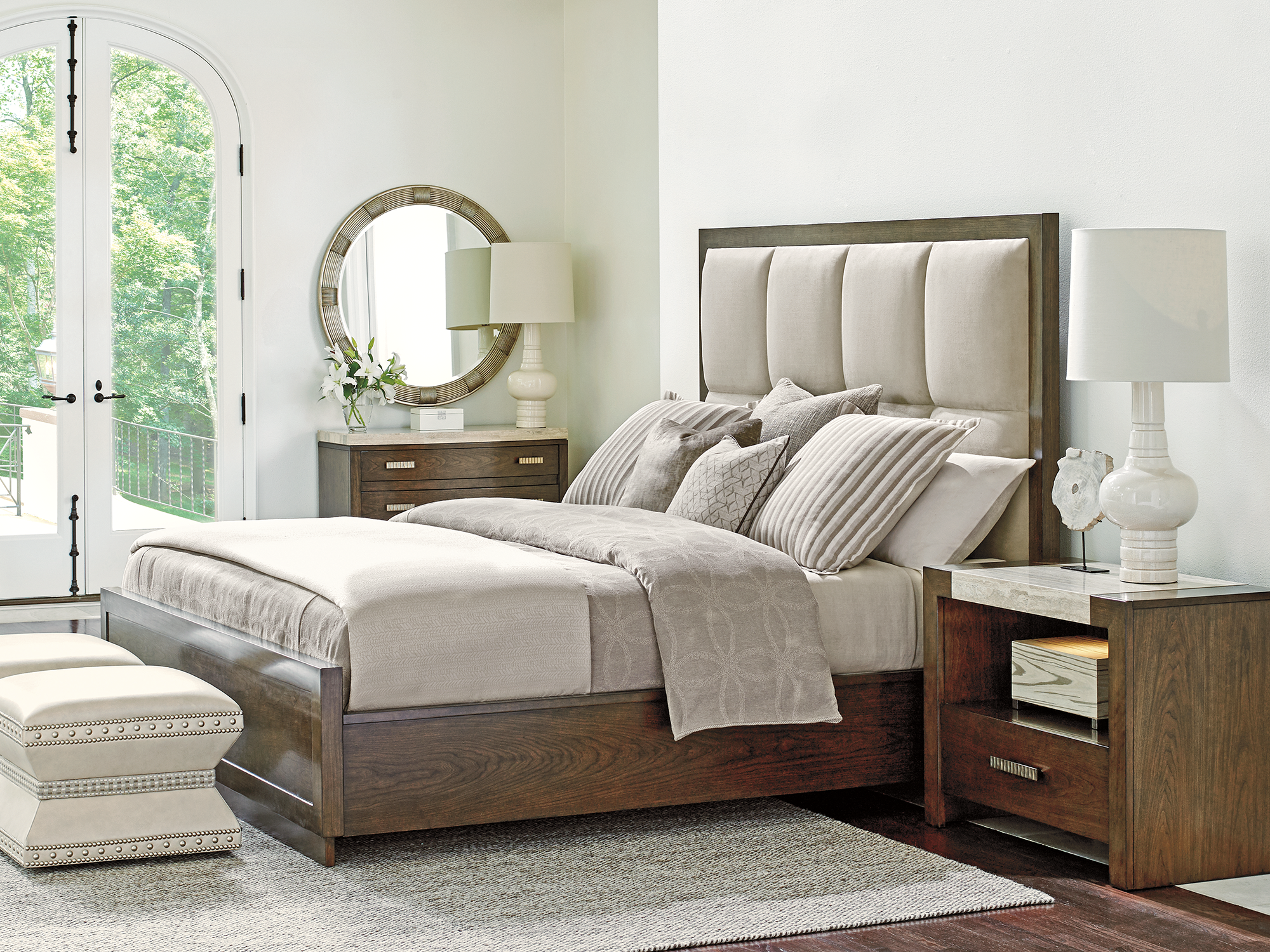 Lexington Laurel Canyon — Transitional bedroom featuring a wood-frame bed with a channeled upholstered headboard. Paired with stone-top nightstands, nailhead-trimmed ottomans, and a sunburst mirror in a bright, arched-window setting.