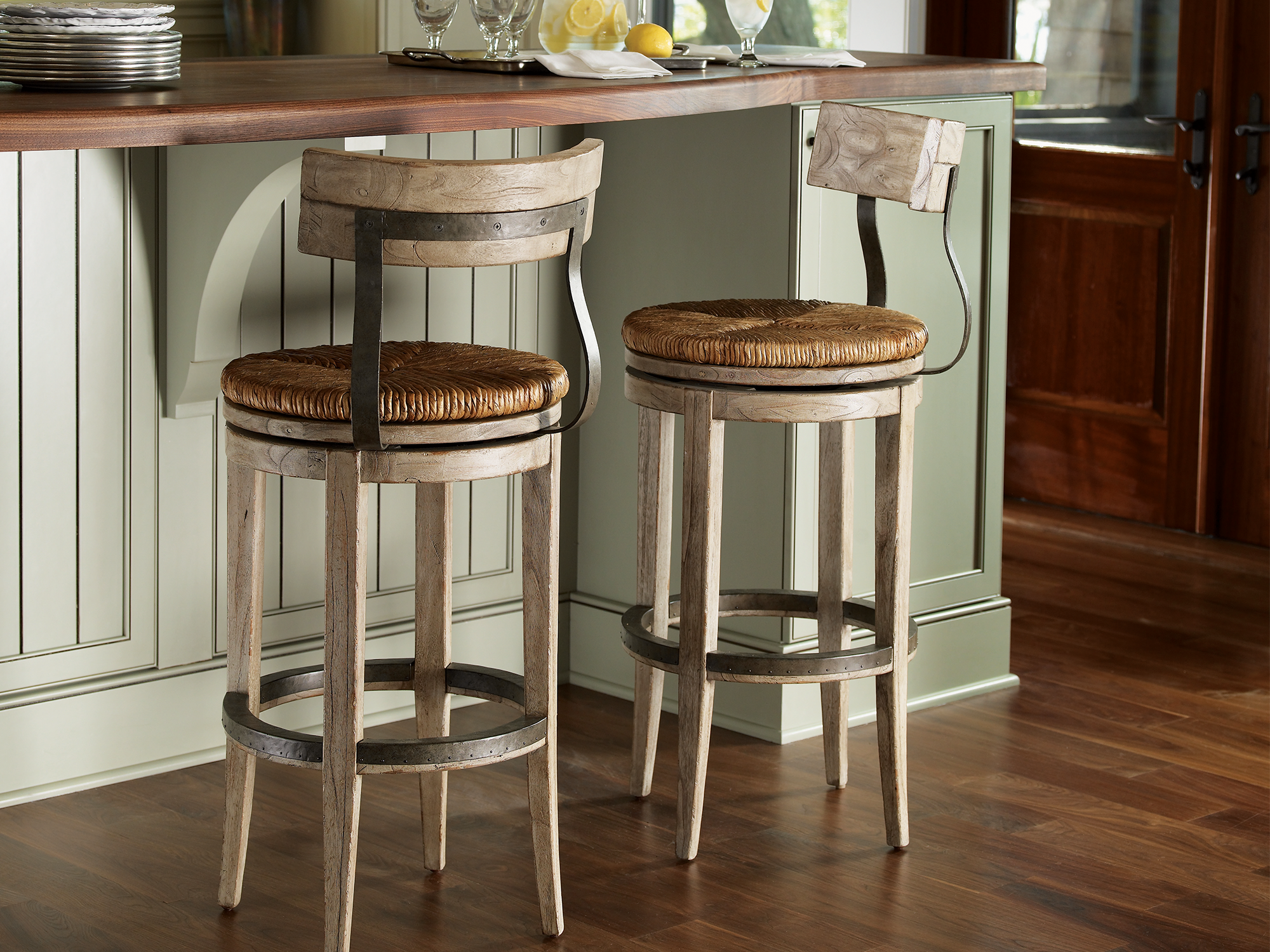 Lexington Twilight Bay — Swivel counter stools with woven rush seats and a rustic driftwood finish. Featuring industrial metal accents and a curved backrest, these stools are set against a sage green kitchen island for a refined coastal look.