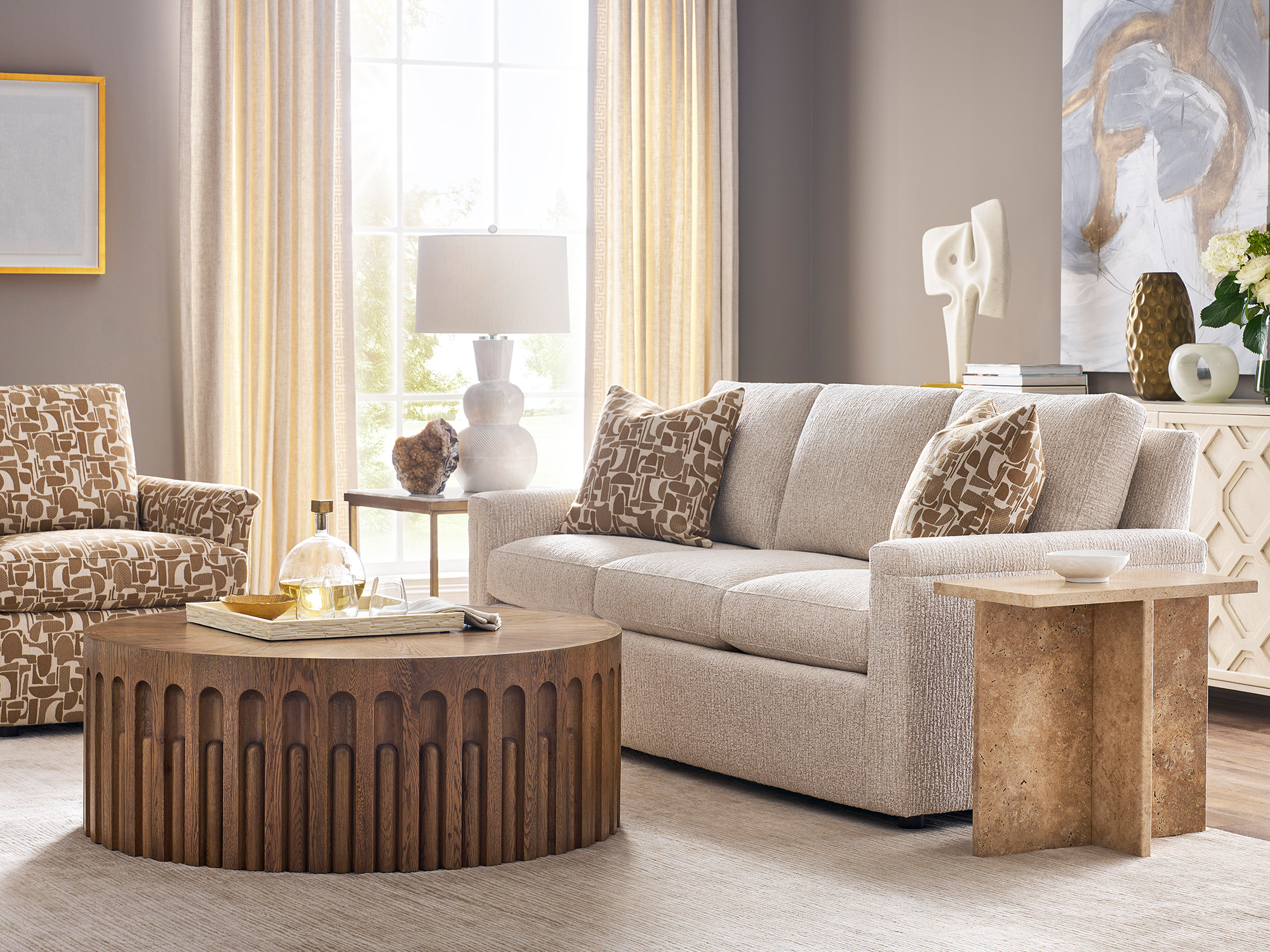 Sherrill 48 Series — modern transitional living room featuring a neutral textured track-arm sofa, a drum-style wooden coffee table with a fluted architectural base, and an abstract-patterned accent chair in a sophisticated, sun-filled setting.