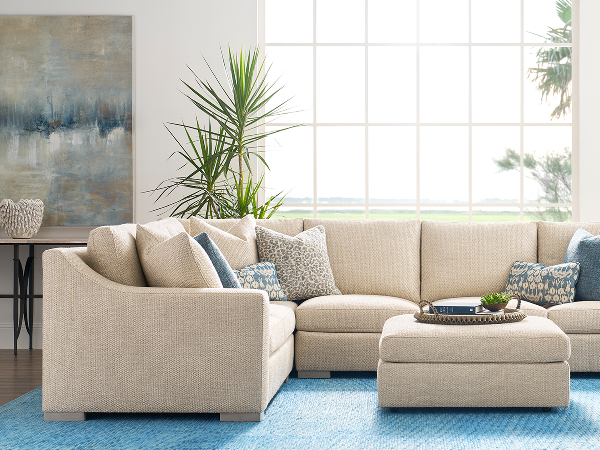 Sherrill 6 Series — airy coastal living room featuring a neutral textured sectional with sloped arms, a matching square cocktail ottoman, and vibrant blue-toned accent pillows on a bright azure area rug, set against a backdrop of floor-to-ceiling windows.