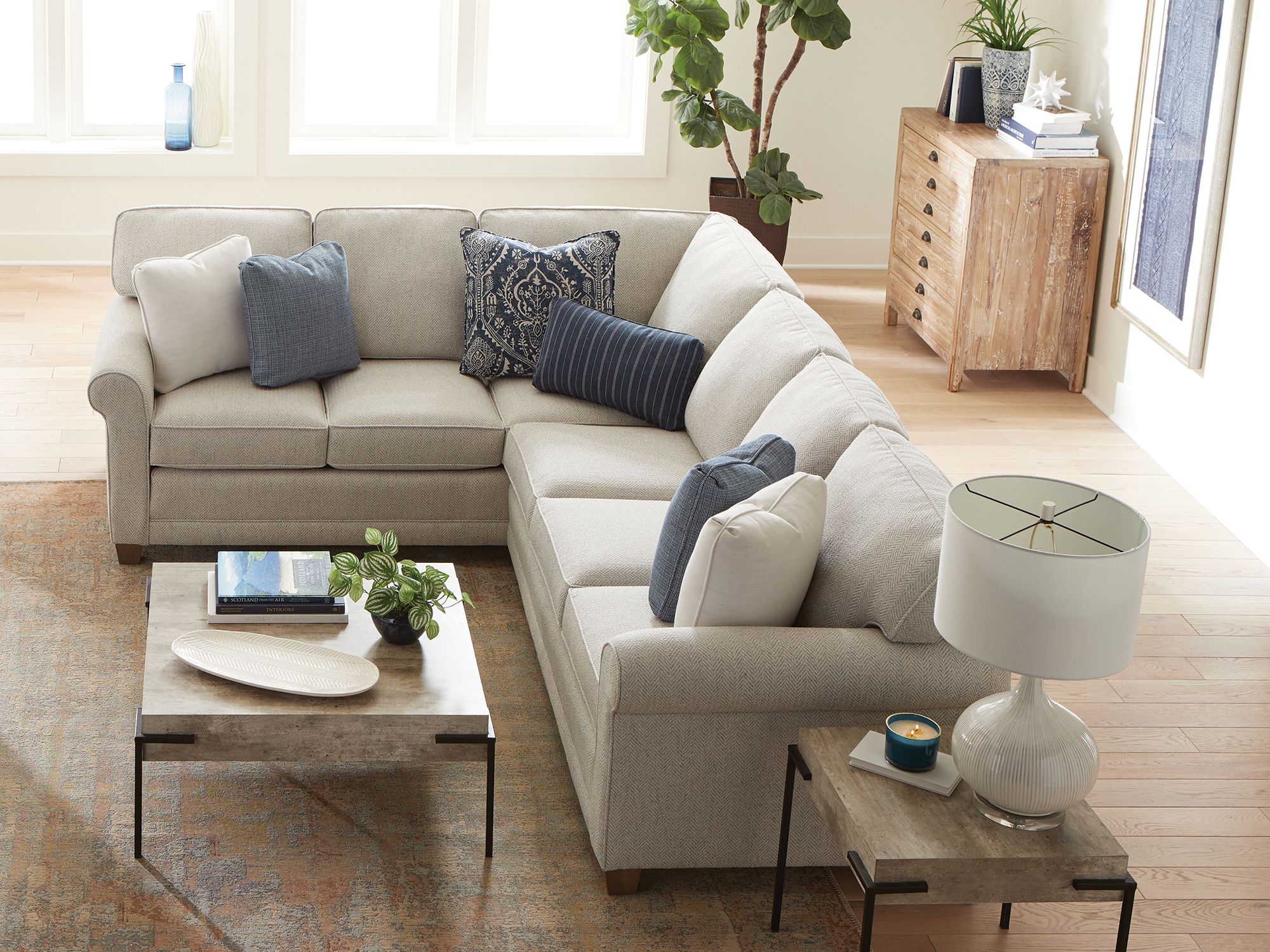 Smith Brothers of Berne — casual L-shaped sectional upholstered in a light grey herringbone fabric with rolled arms, accented by navy and cream patterned toss pillows and paired with a weathered-wood coffee table on a black metal frame.