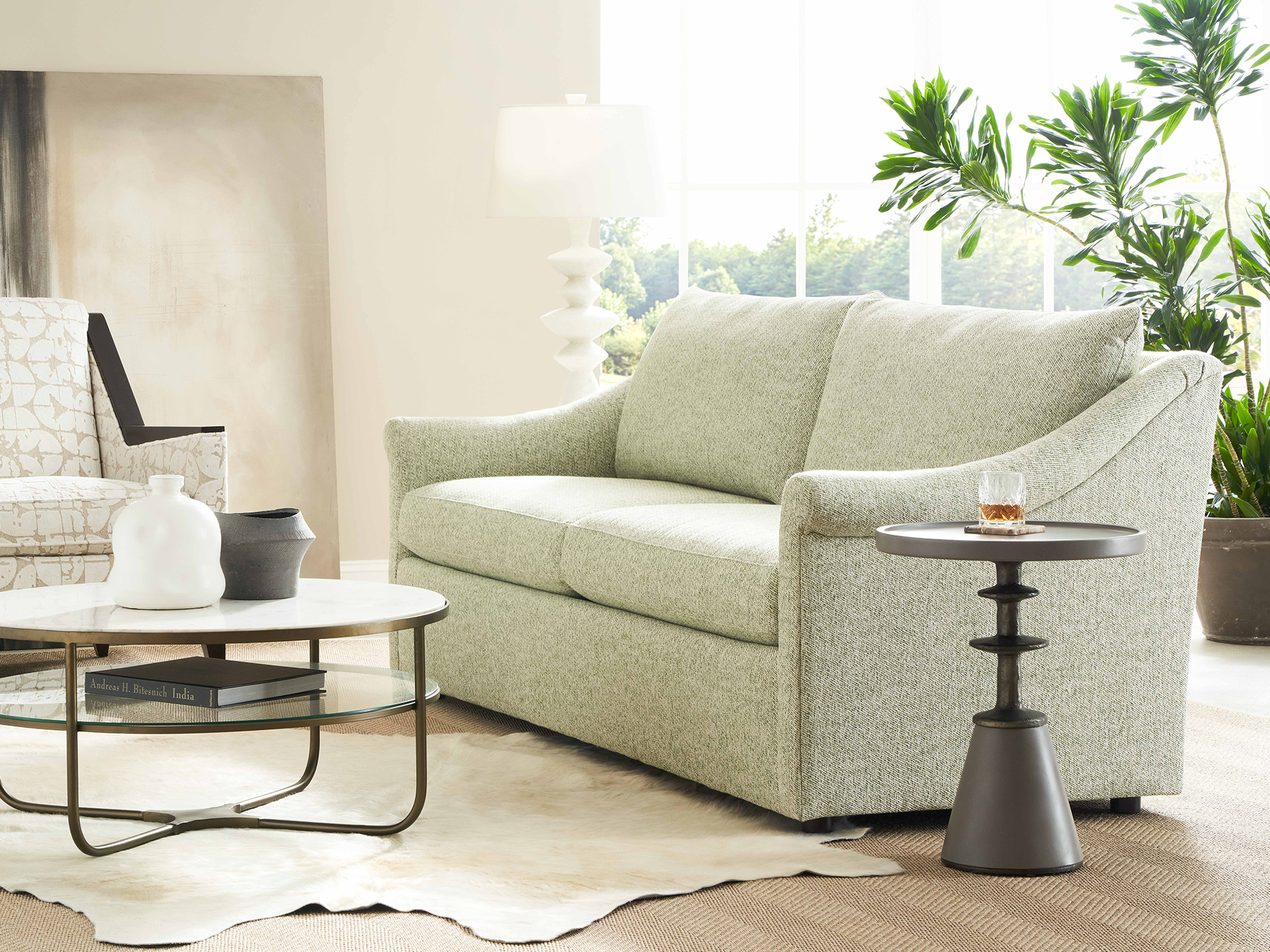 Sherrill 42 Series — transitional living room featuring a pale sage green textured sofa with flared arms, a round marble-top cocktail table with a glass lower shelf, and a unique sculptural pedestal end table on a layered hide and jute rug.