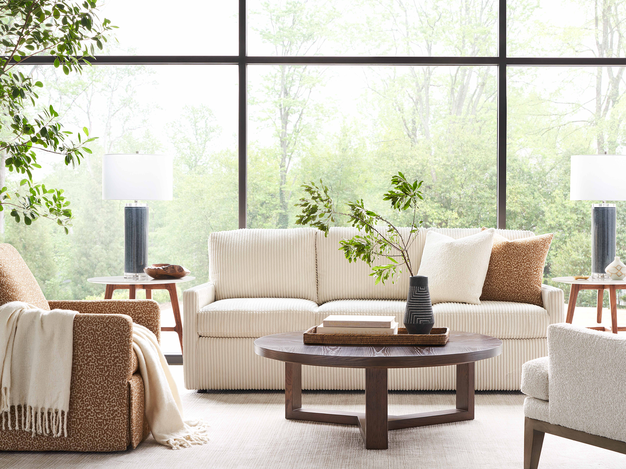 Sherrill 43 Series — modern transitional living room featuring a cream ribbed-fabric sofa with a dressmaker skirt, a dark wood round coffee table, and patterned accent chairs in an airy space with floor-to-ceiling windows.